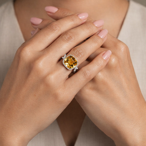 Close-up of a woman's hand wearing a ring with a yellow citrine gemstone and diamonds, against a neutral background.