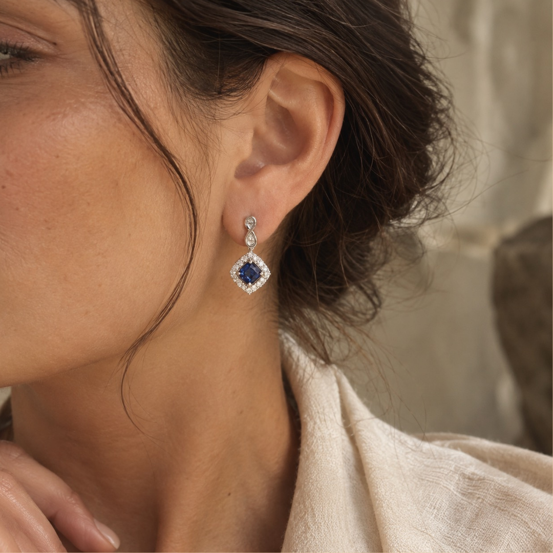 A close-up of a woman's ear wearing a diamond-shaped drop earring with a deep blue sapphire center stone and a halo of small clear stones in a white gold setting.