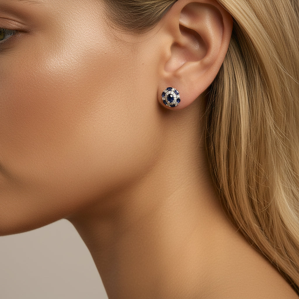 Close-up of Clair de Saphir vintage-style stud earring worn on a model’s earlobe, showing the blue sapphire and diamond halo, with smooth bronzed tan skin and a few strands of dark hair visible.