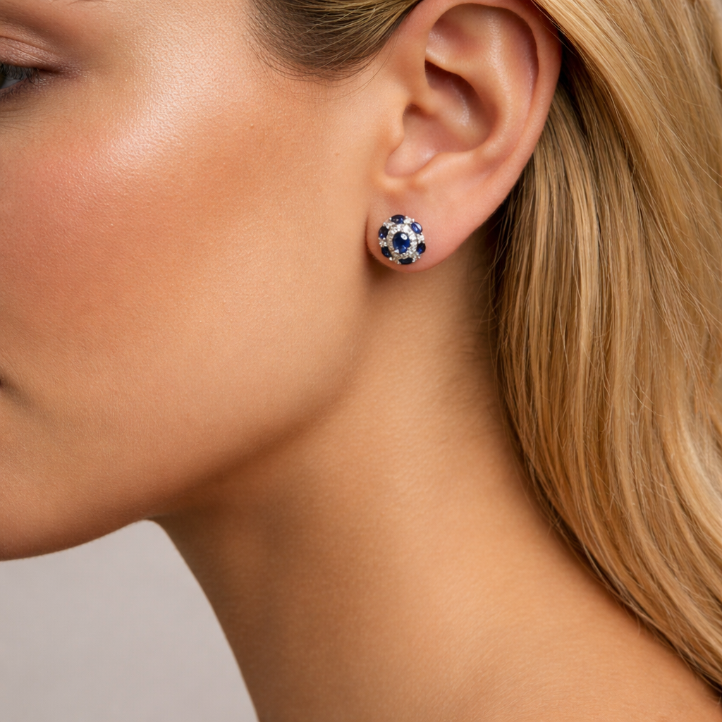 Close-up of Clair de Saphir vintage-style stud earring worn on a model’s earlobe, showing the blue sapphire and diamond halo, with smooth bronzed tan skin and a few strands of dark hair visible.